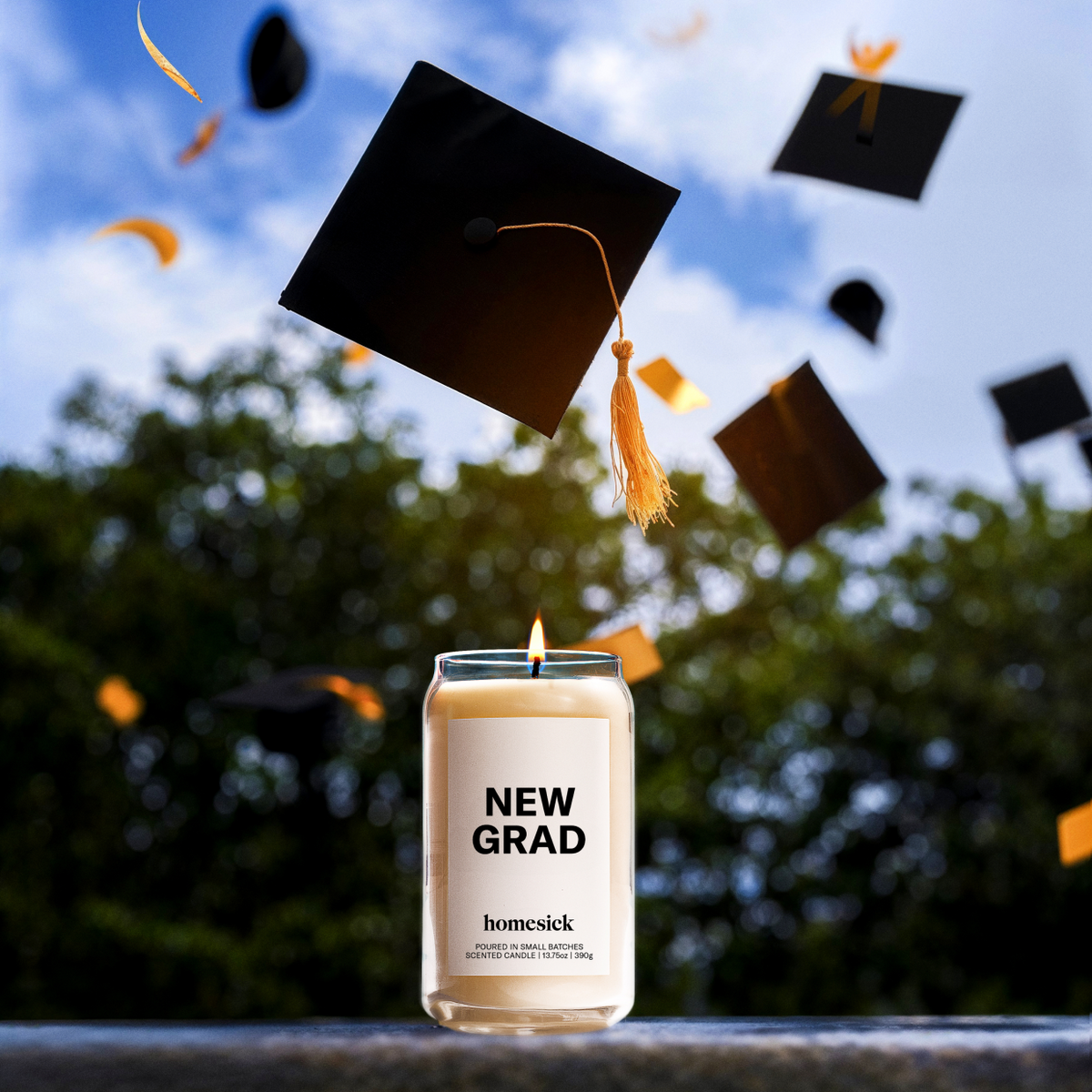 A lifestyle image of the New Grad Homesick candle in an outdoor environment surrounded by falling graduation caps.