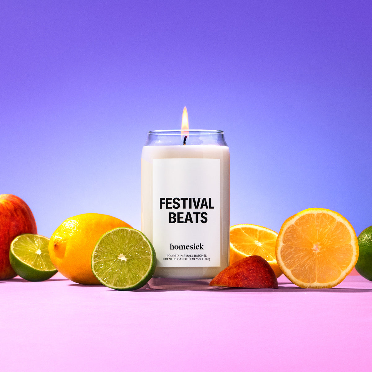 A lit Festival Beats Homesick candle on a colorful background surrounded by various fruits