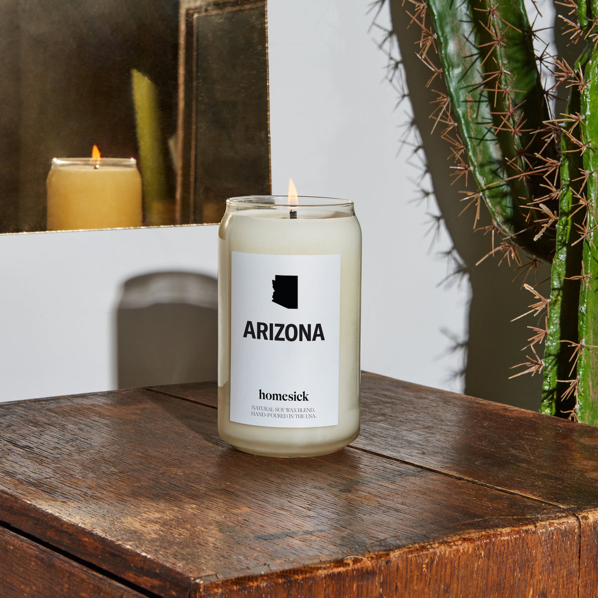 A close up of a lit Arizona candle on a wooden table. In the corner, one can see a cactus. 