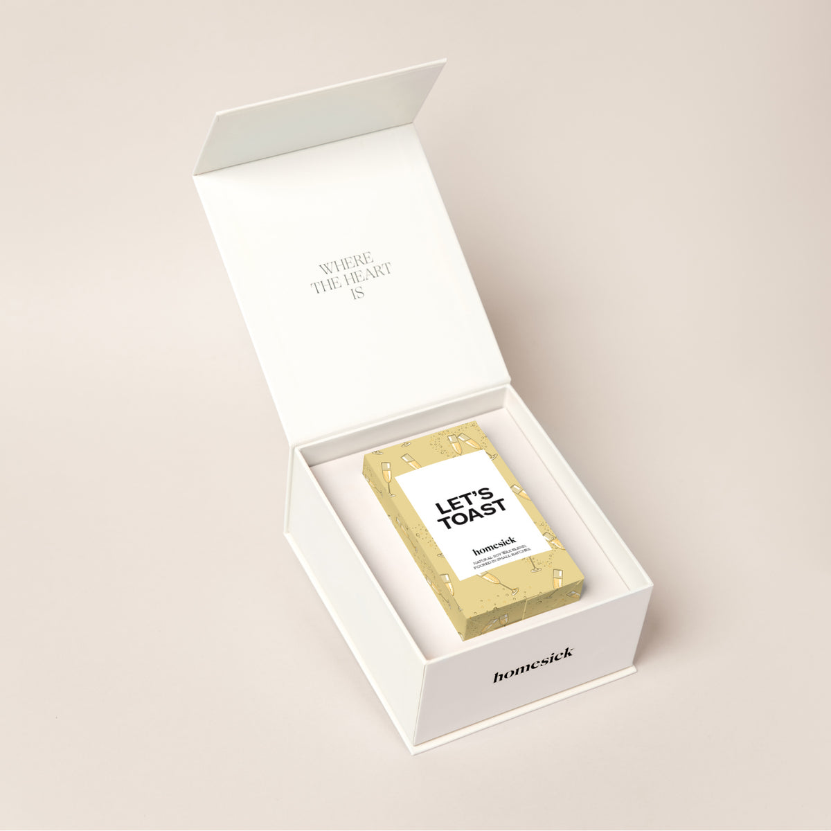 A white candle box that displays how a candle fits perfectly inside. The top lid packaging reads, "where the heart is" and inside is the Let's Toast candle. 