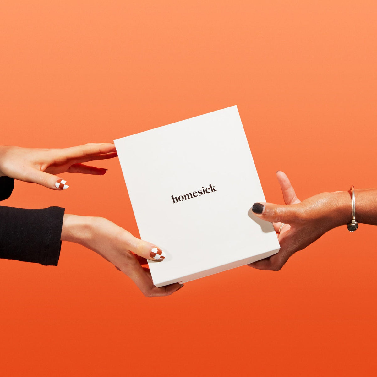 Two model hands holding the Homesick Candle box in front of red orange background.