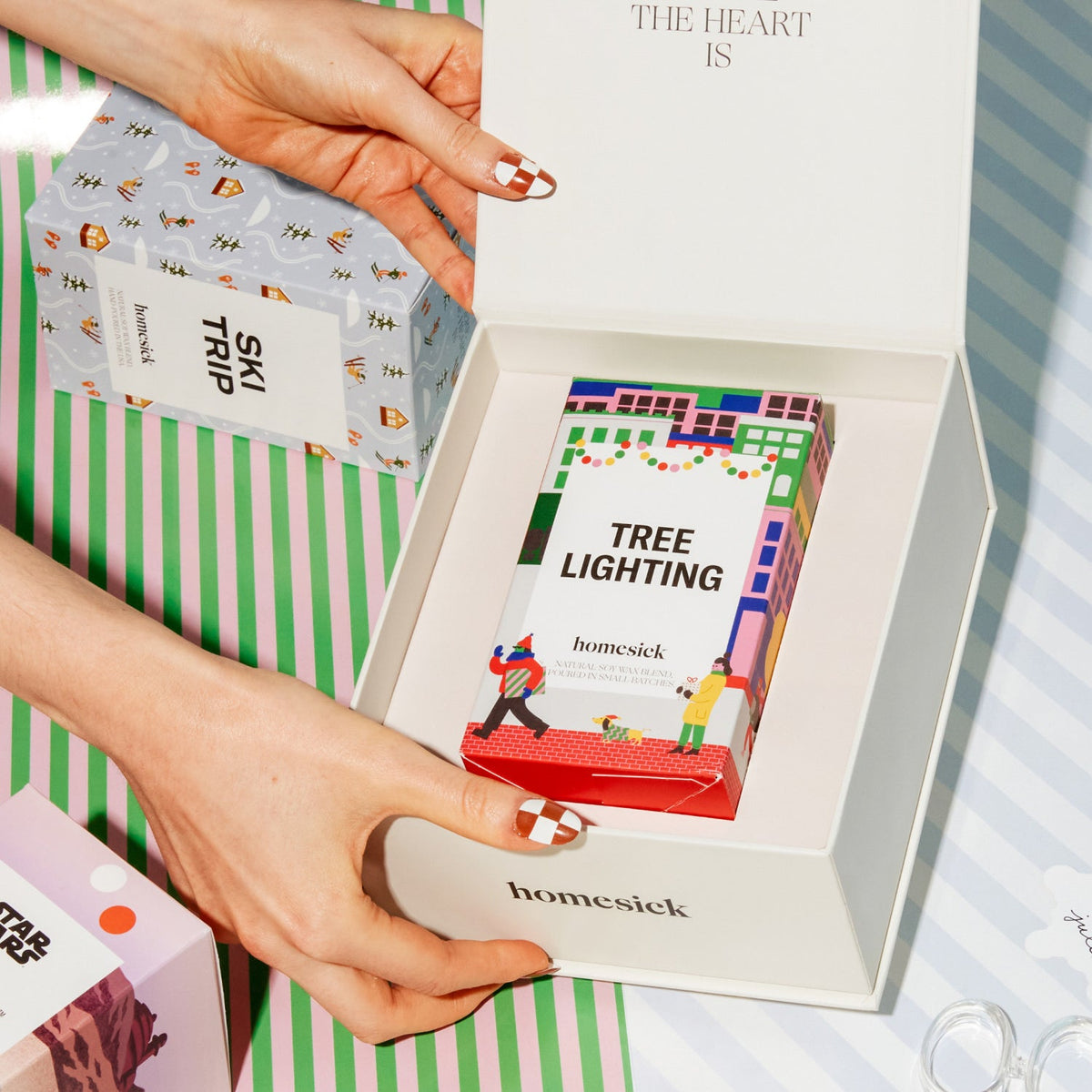 Two hands showing the Homesick candle box open with the Tree Lighting Candle inside. Behind the candle box is a background of wrapping paper print.