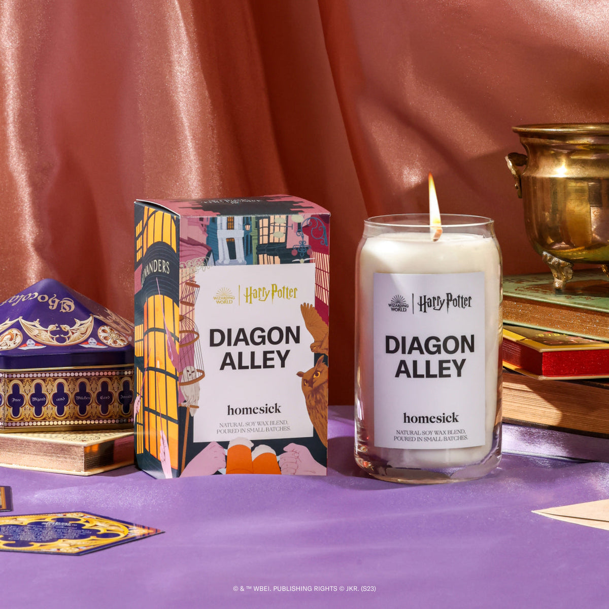 A stylized shot of the Harry Potter Diagon Alley candle and its packaging alongside various fantasy props that would fit the series. The surface is a silky purple fabric and background is a silky salmon fabric.