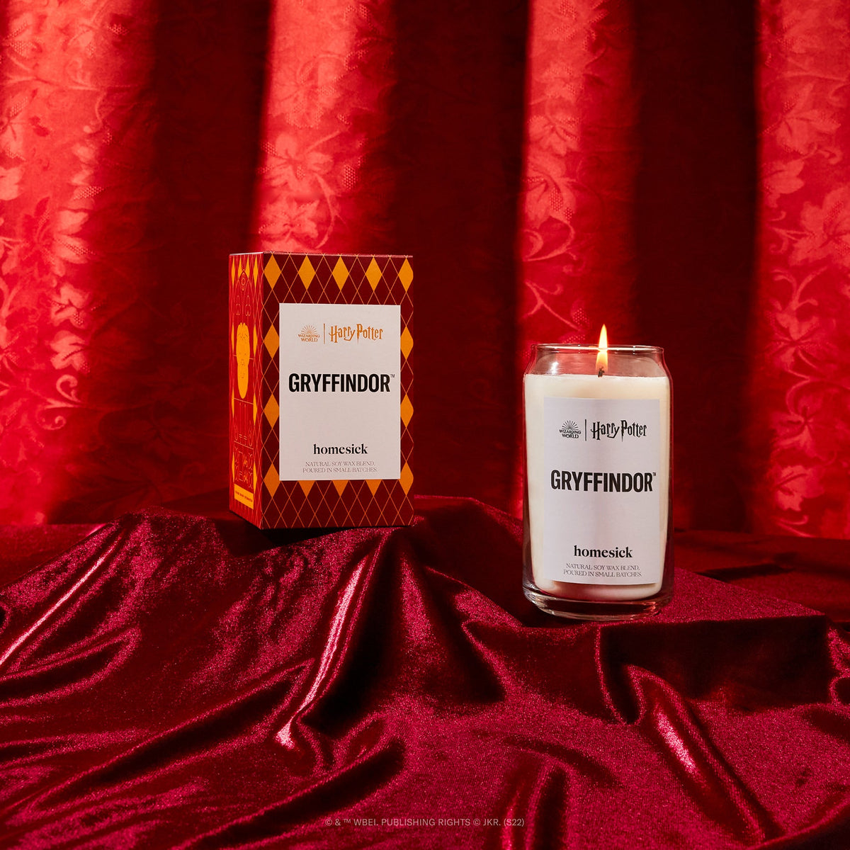 A side by side image of the Gryffindor candle and its packaging. Shot in a very moody red environment over a metallic red surface and a red pattern fabric as the background.