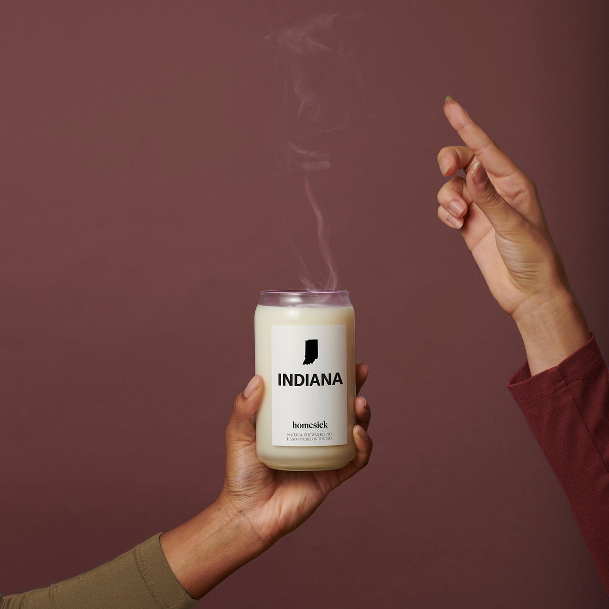A hand holding the Indiana Homesick candle that has just been unlit by someone and their fingers.