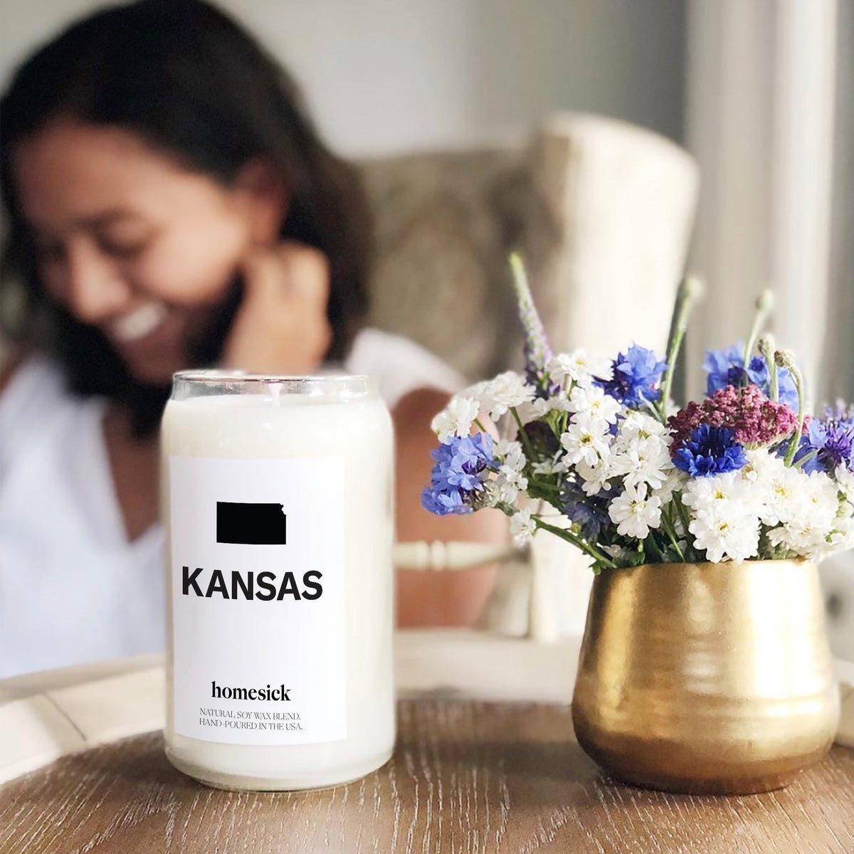 A close up shot of a Kansas Homesick candle on the left and a small flower arrangement in a gold pot on the right. The background is blurry but there is a woman sitting in a chair.