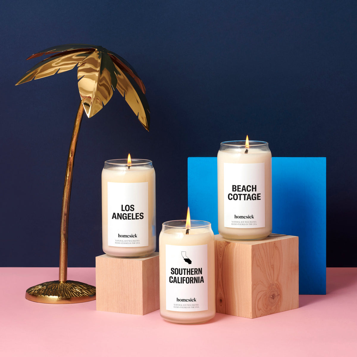 A group shot of the California Coast inspired candles at Homesick. The SoCal candle is on the pink surface with the LA candle and Beach Cottage candle above it on wooden cube pedestals. On the left is a small gold palm tree for decor with a navy blue background.