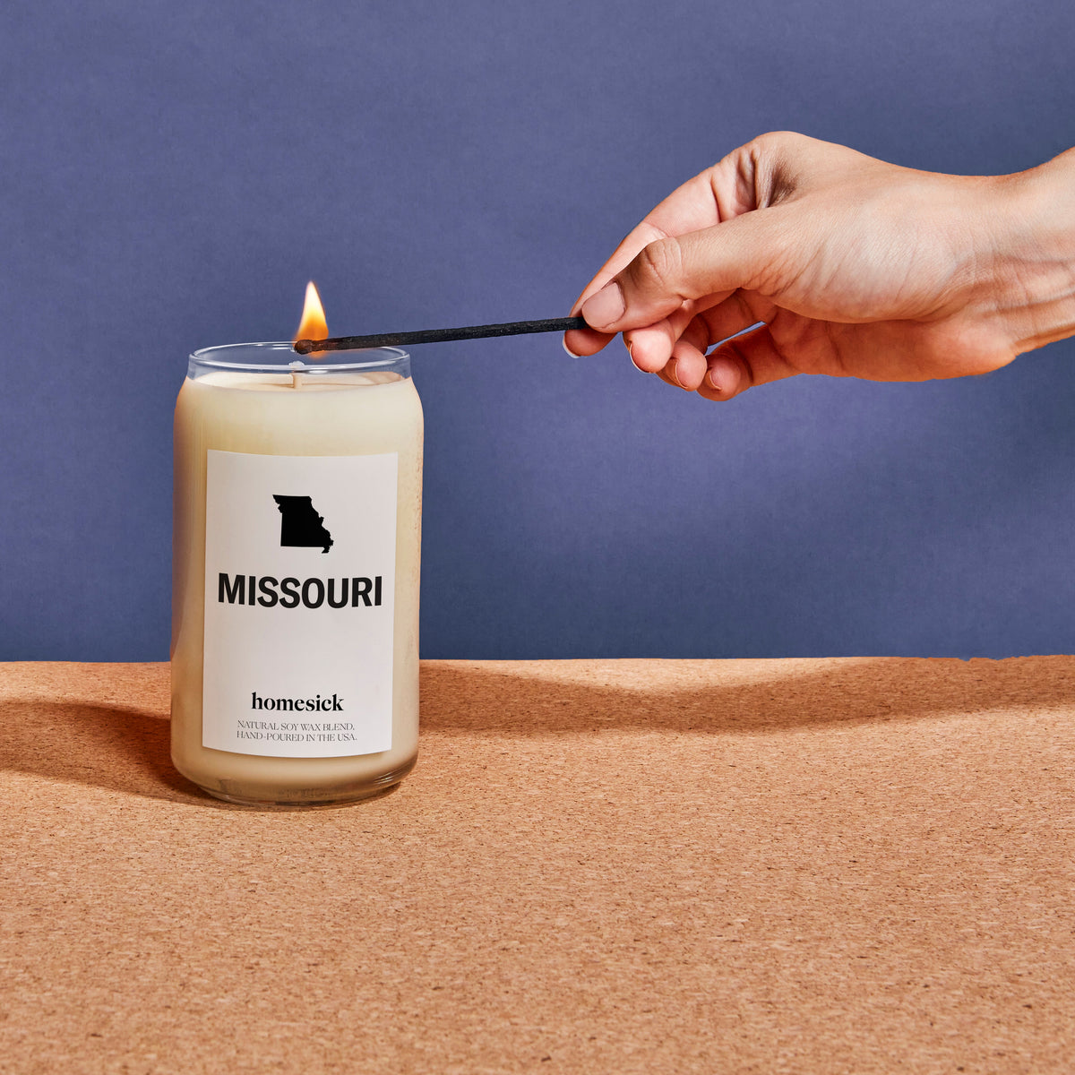 A close up of a Missouri candle displayed on a cork board surface with a navy background. There is a hand entering from the left side that is using a match to light the candle.