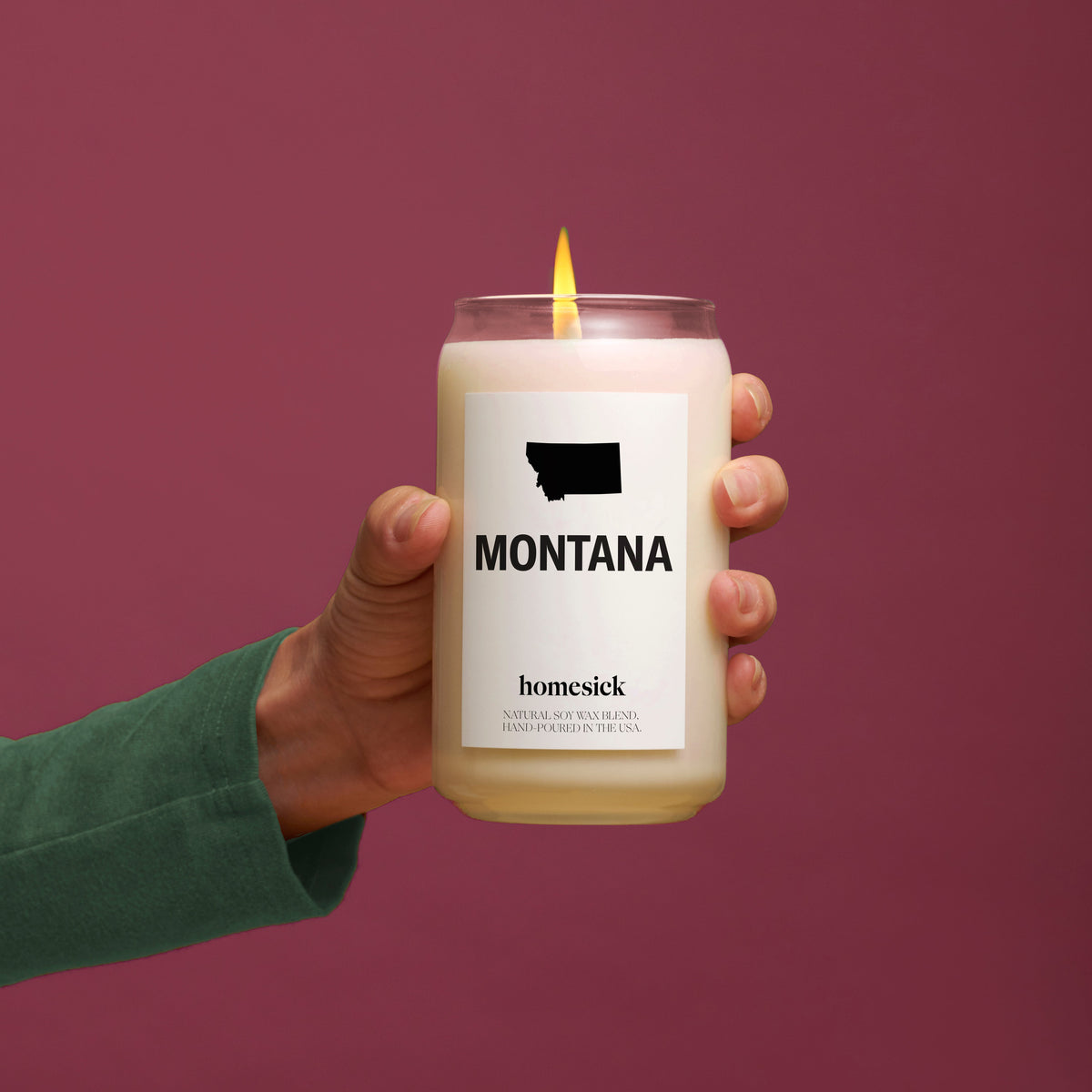A hand holding a lit Montana candle in front of a wine background.