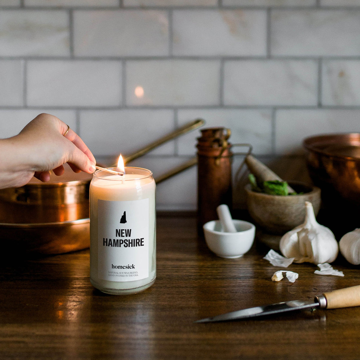 A hand going to light the New Hampshire candle with a match inside a kitchen that has various prep items.