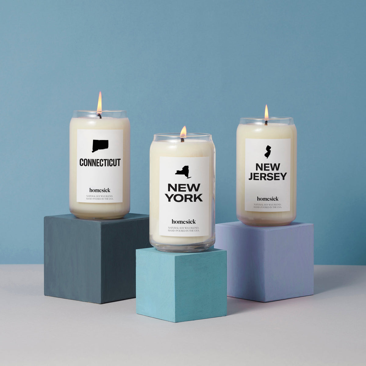 A group product shot of the Northeast inspired candles offered at Homesick. Each are displayed on cube pedestal but each are a different shade of blue. The candles shown are Connecticut, New York and New Jersey.