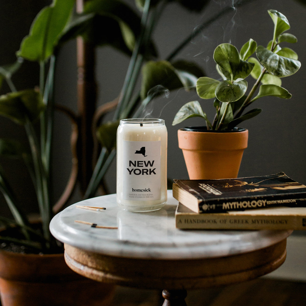 The New York Candle displayed on a rounded marble table that has various books, matches and potted plants also on the table.